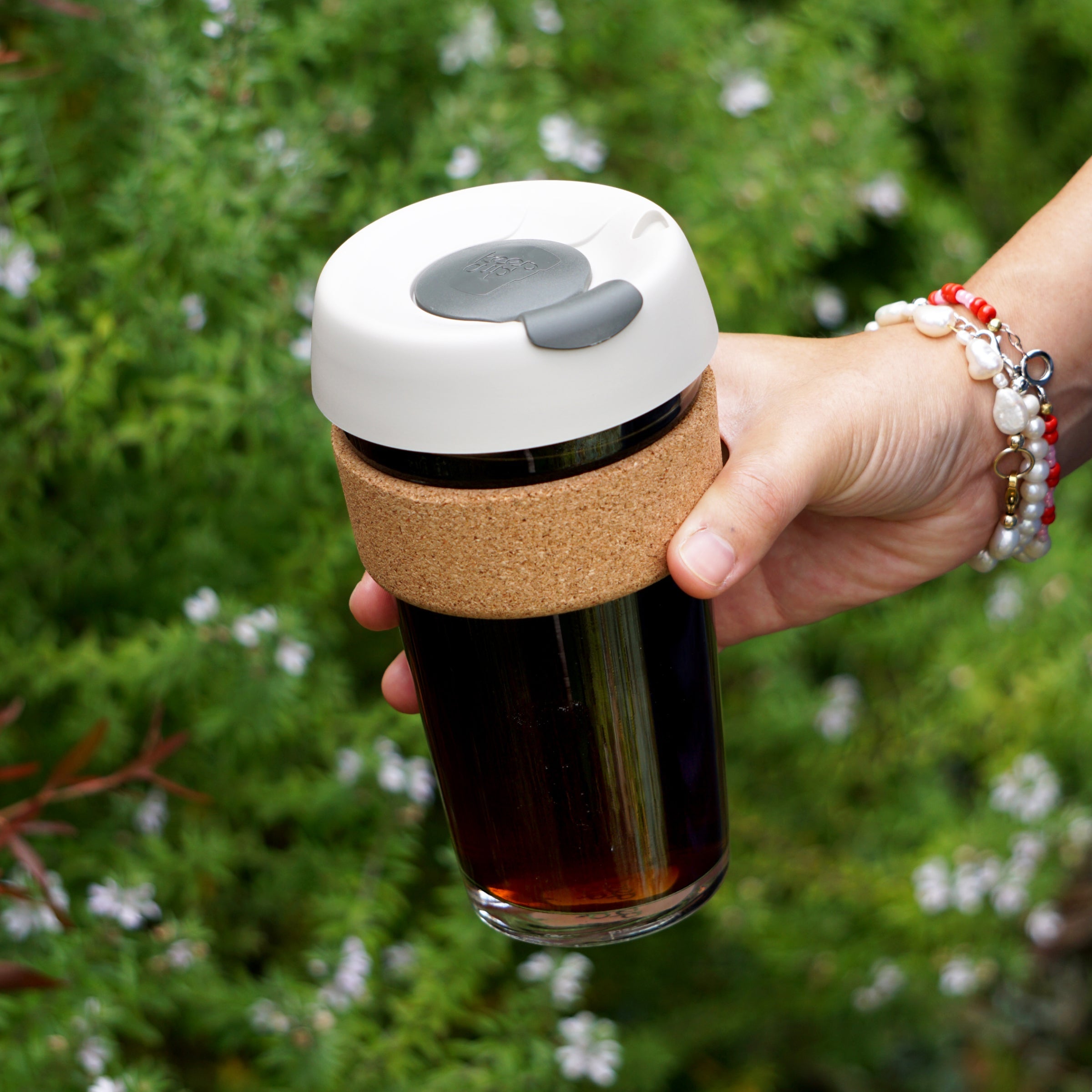 Hand holding a KeepCup Brew Cork L 16oz in Filter, featuring a clear glass cup filled with dark coffee, natural cork band, and white lid with gray plug, against a lush green garden background.