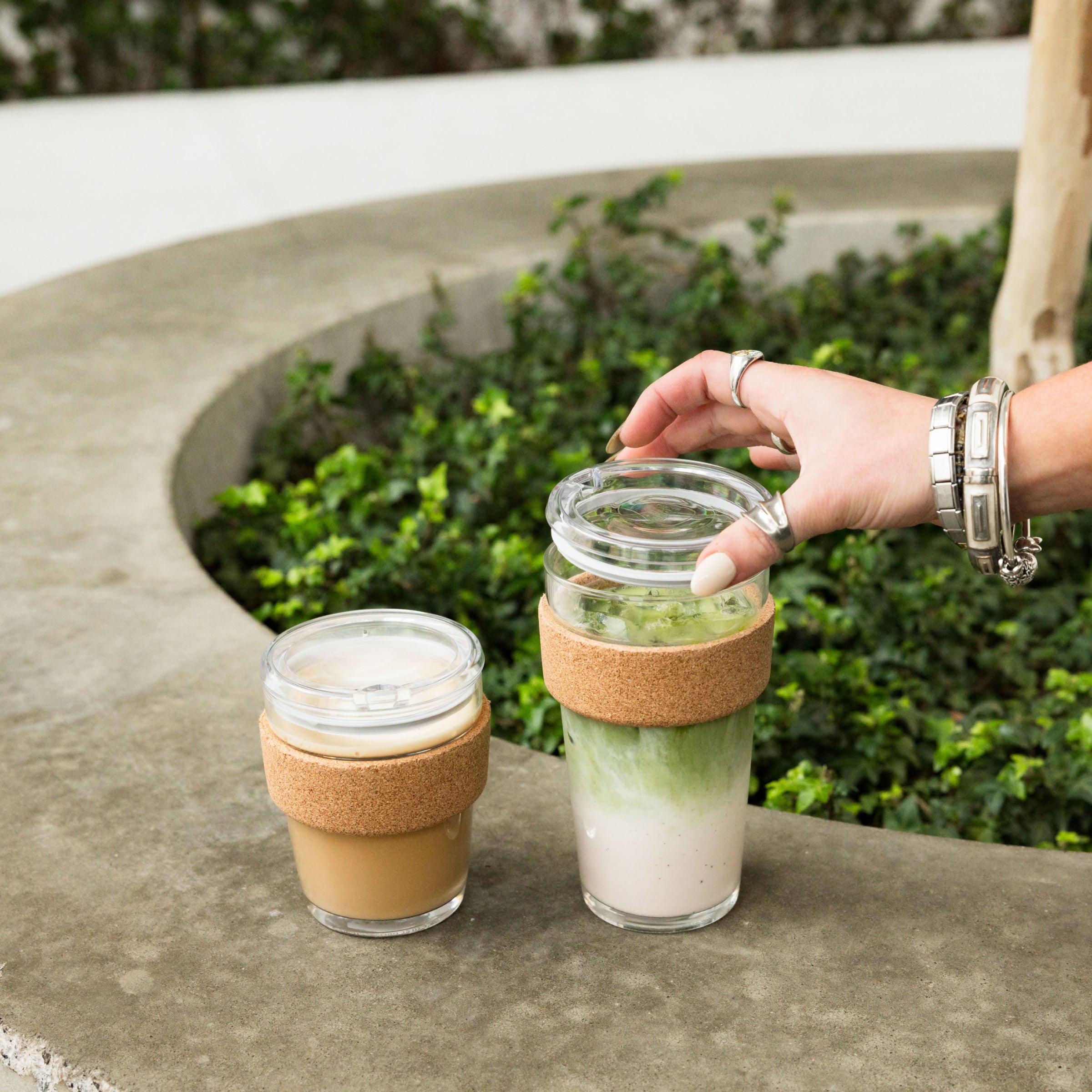 A hand with silver jewelry reaches for the lid of a Brew Cork Quicksip L 16oz KeepCup filled with an iced matcha latte, sitting on a curved concrete ledge beside a smaller cork-banded glass cup containing an iced coffee, with green foliage in the background.