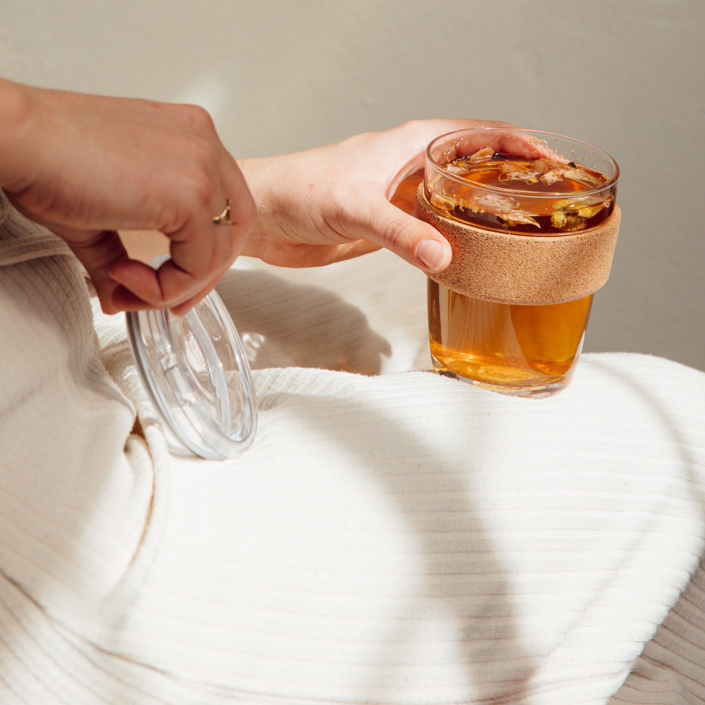 A hand holds a KeepCup Brew Cork Quicksip M 12oz filled with amber-colored tea, with the clear Quicksip lid removed and held in the other hand, set against a soft neutral background with natural lighting.
