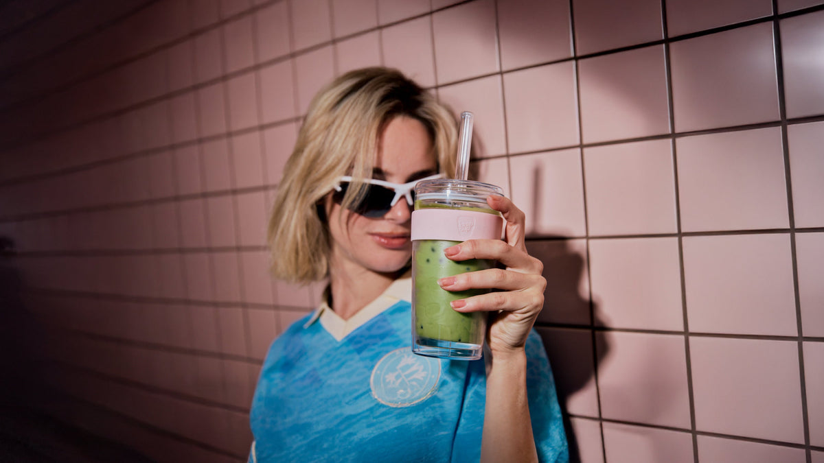 Woman in blue shirt drinking green smoothie from glass tumbler with straw against pink tile wall