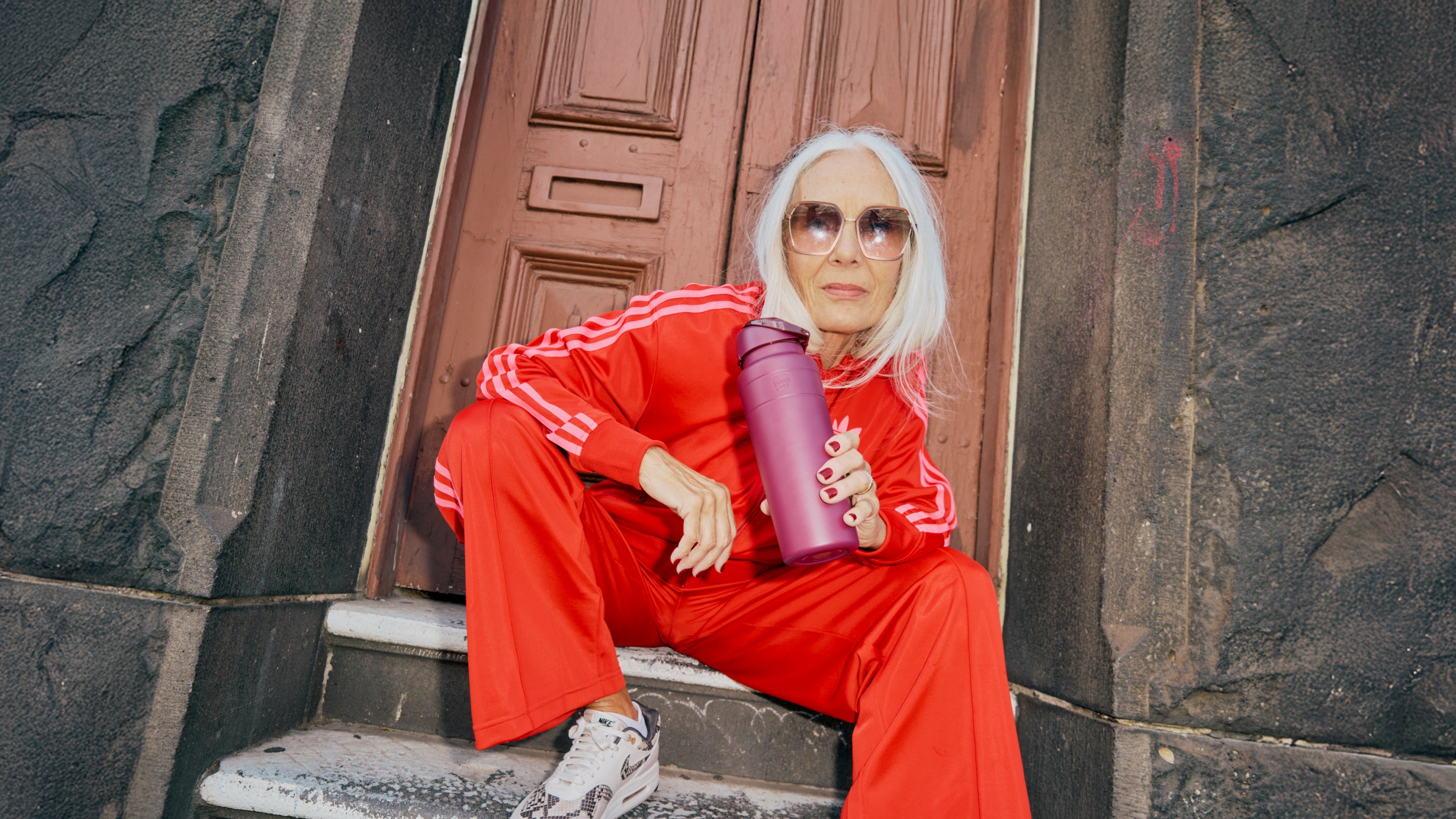 Stylish woman in red tracksuit sitting on steps holding purple insulated water bottle, wearing sunglasses and white sneakers