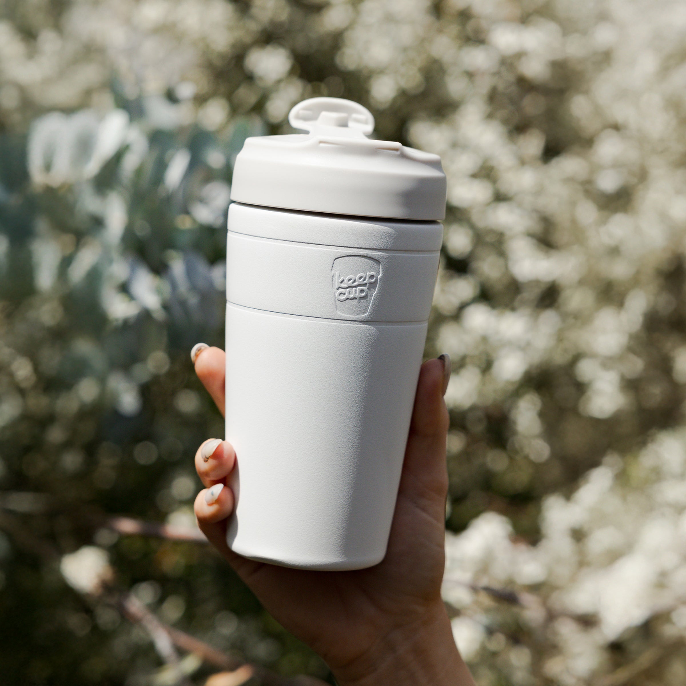 Person holding white KeepCup reusable coffee cup with lid outdoors against blurred green foliage background
