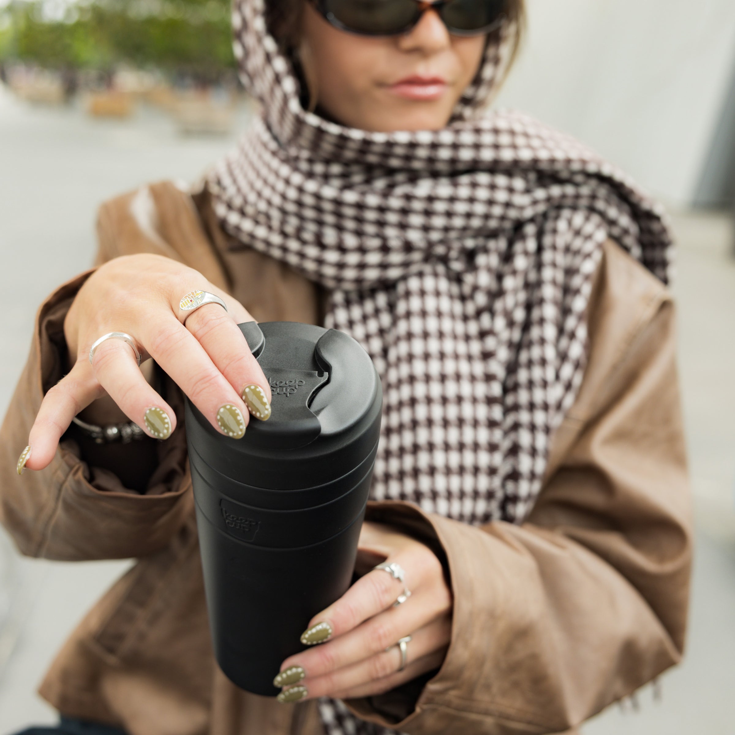 A woman wearing sunglasses, a brown jacket, and a checkered scarf holds a KeepCup Commuter XL 20oz in Black, her olive-green manicured nails resting on the black lid as she prepares to take a sip outdoors.