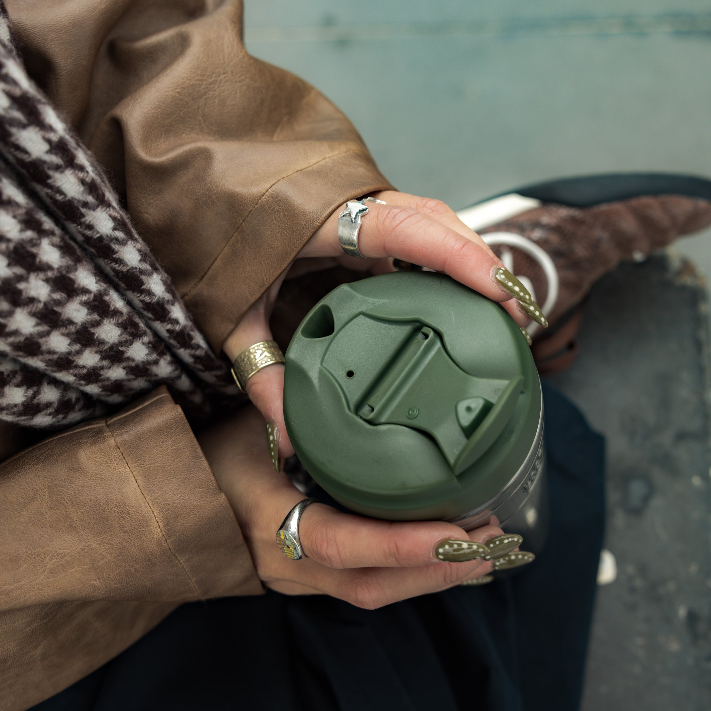 Person holding sage green reusable travel coffee cup with lid, wearing brown coat and gold rings