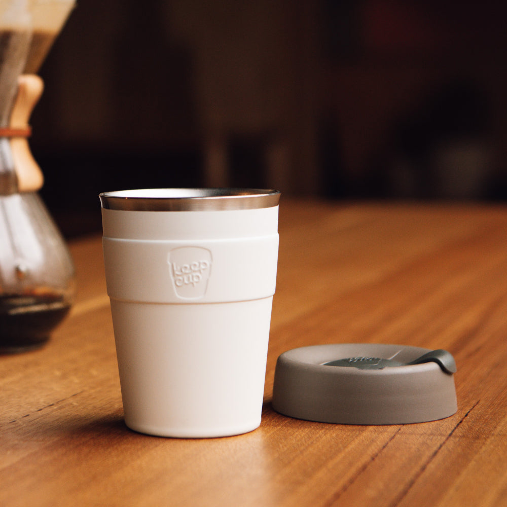 White KeepCup reusable coffee cup with gray lid sitting on wooden table in cozy cafe setting