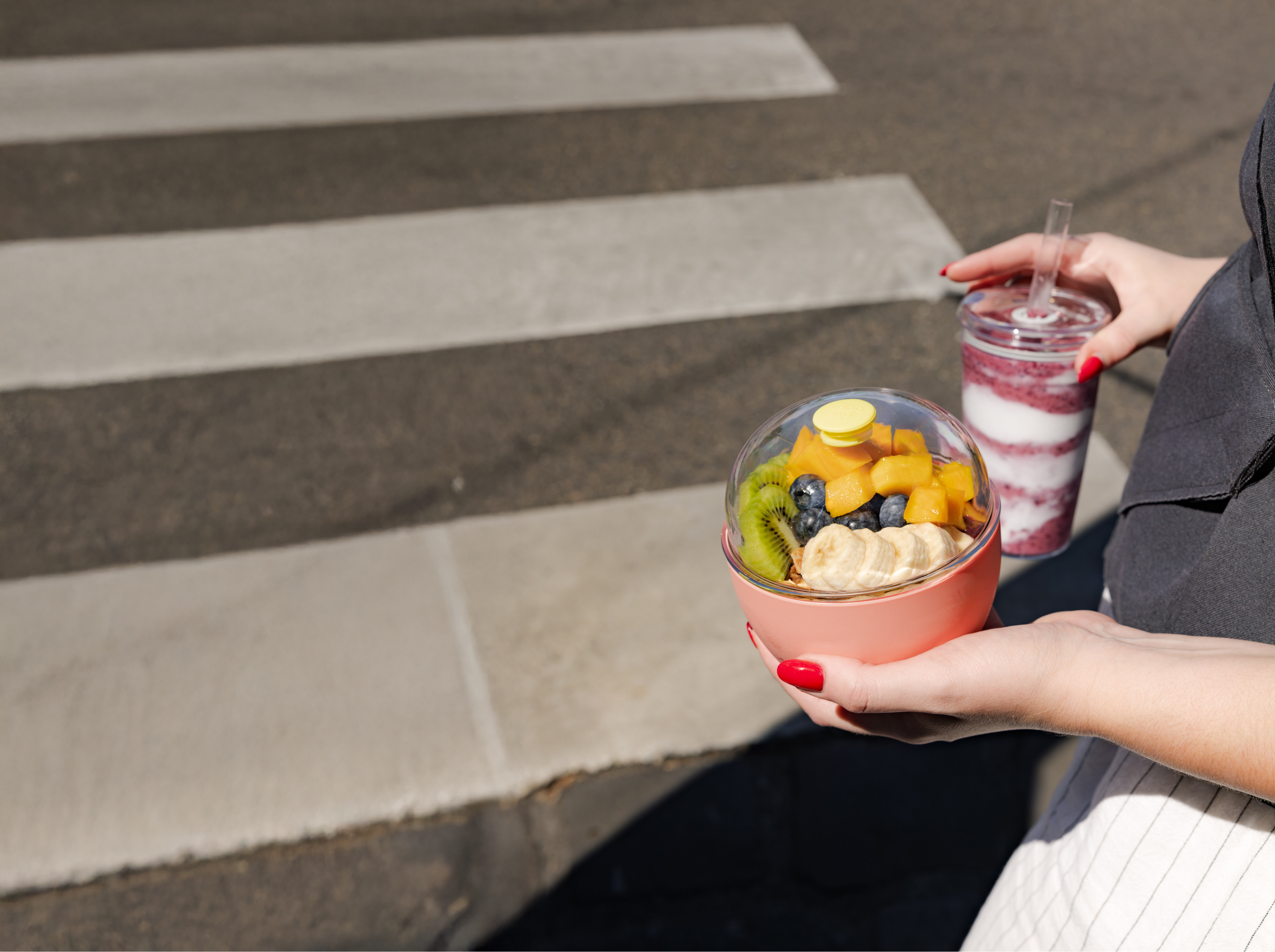 Woman holding pink glass bowl with fresh fruit and berry smoothie, sitting on concrete steps outdoors
