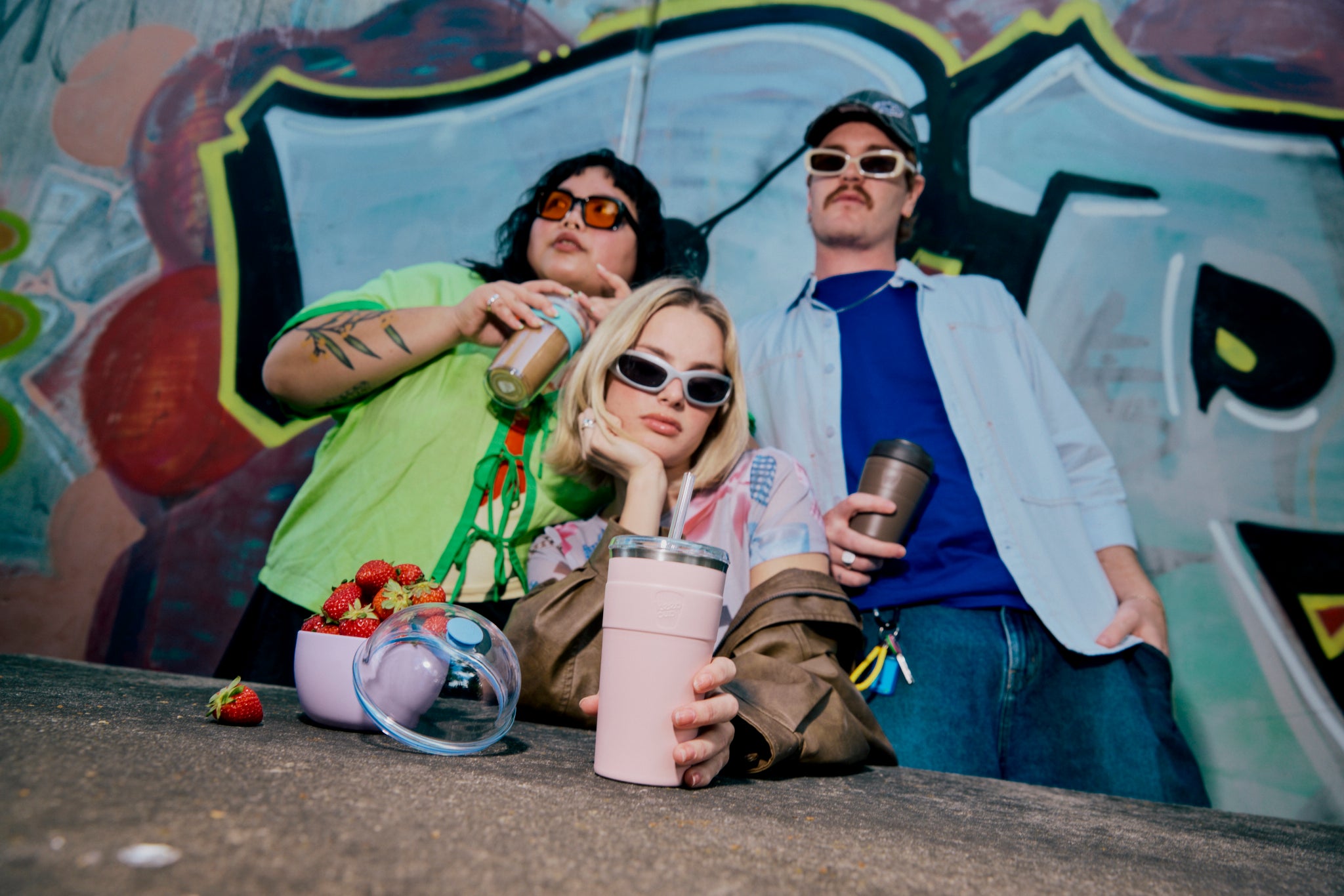 Three friends relaxing against colorful graffiti wall wearing sunglasses, holding reusable drink cups and sharing strawberries from glass bowl