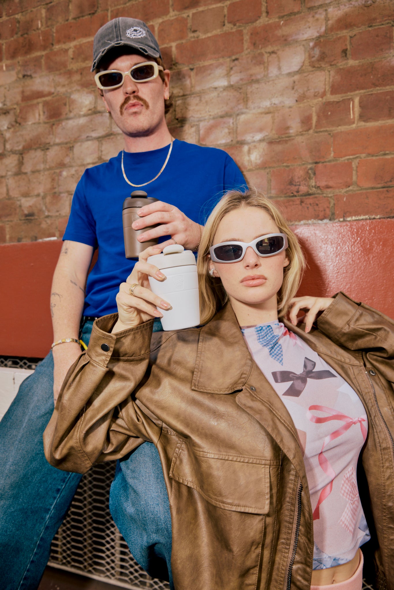 Two people wearing matching white rectangular sunglasses posing against brick wall, woman in brown leather jacket, man in blue t-shirt and cap