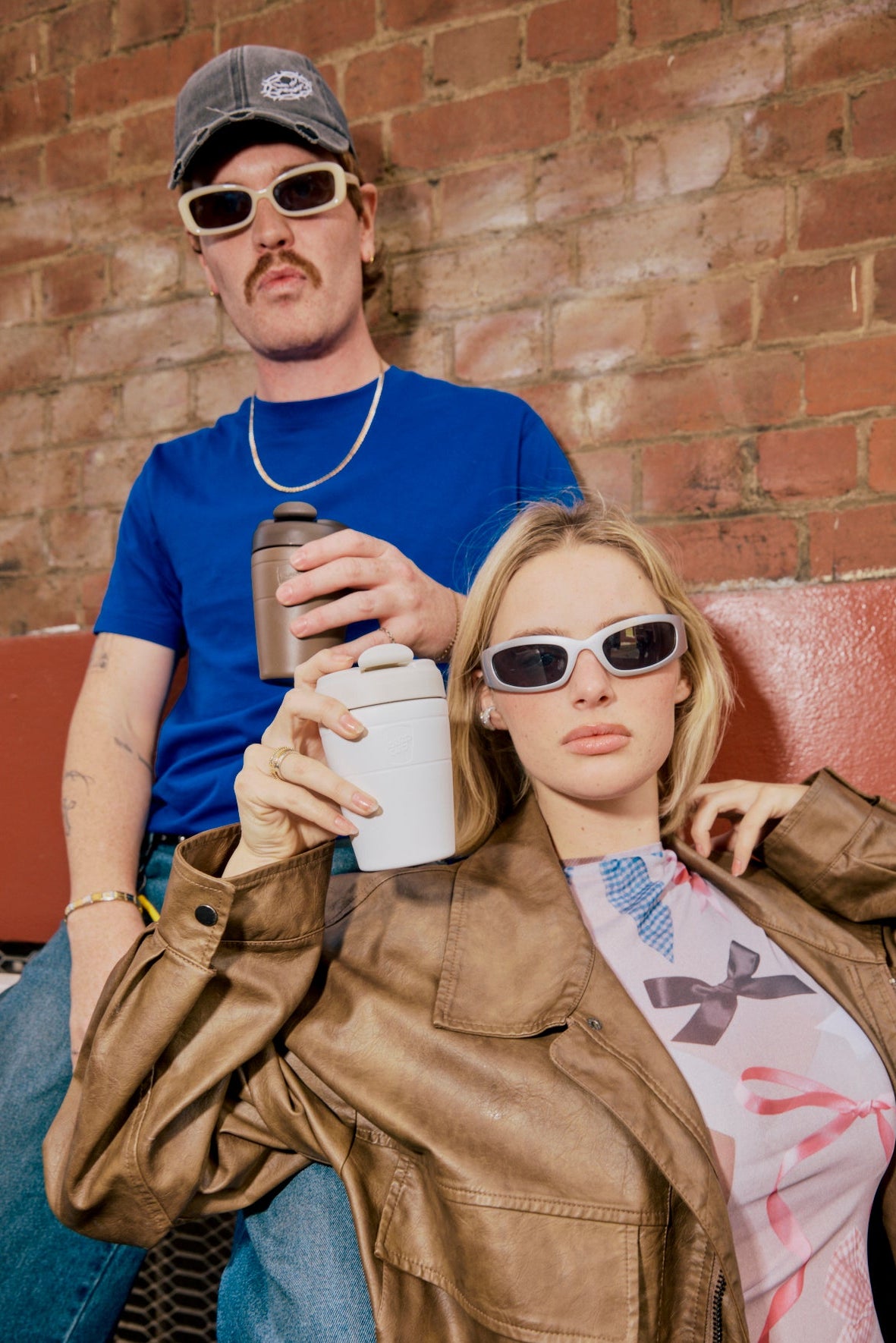 Two people sitting on a couch against a brick wall, holding drinks.