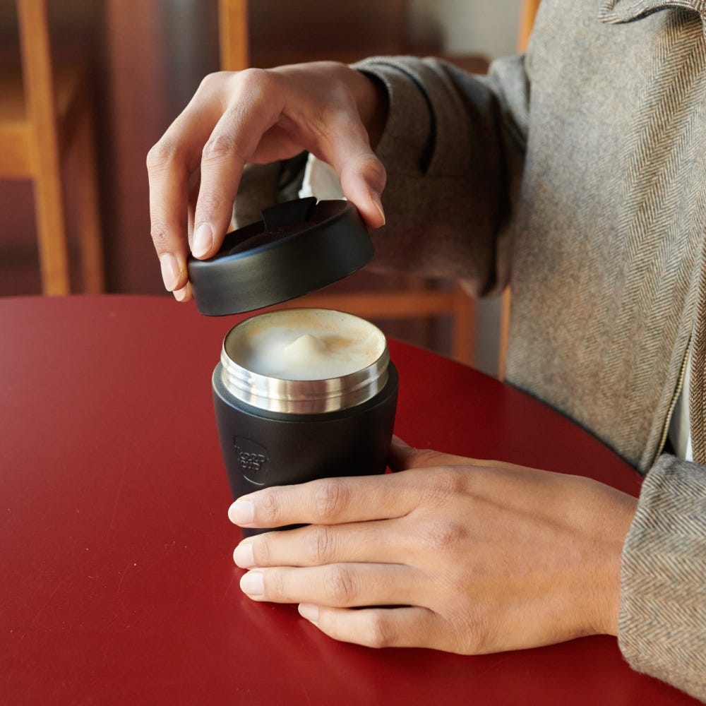 A person in a grey herringbone jacket opens the lid of a KeepCup Traveller S 8oz in Black, revealing a foamy cappuccino inside, while seated at a red café table.