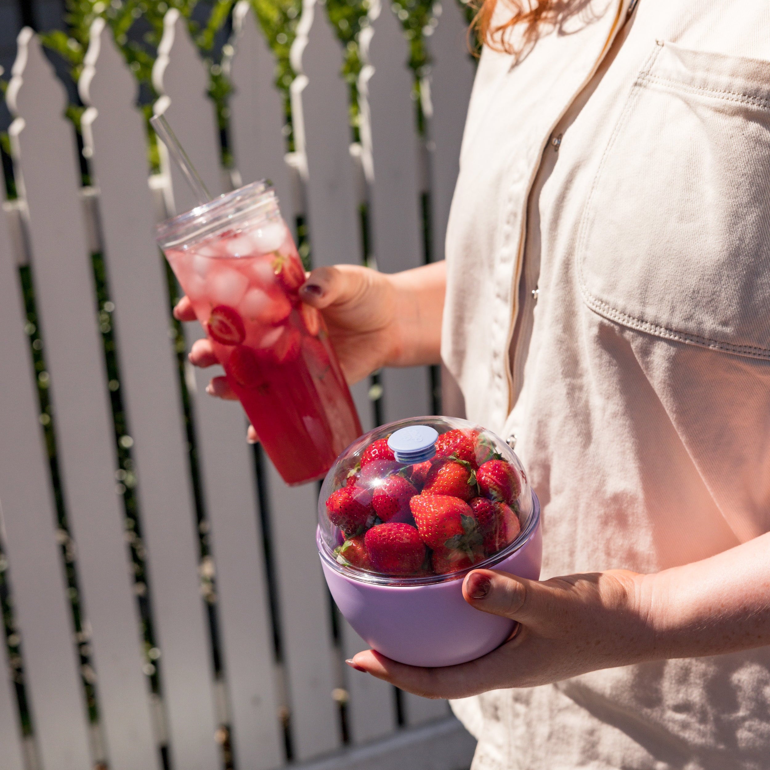 Person holding a tumbler with a strawberry drink and a container of strawberries outdoors.