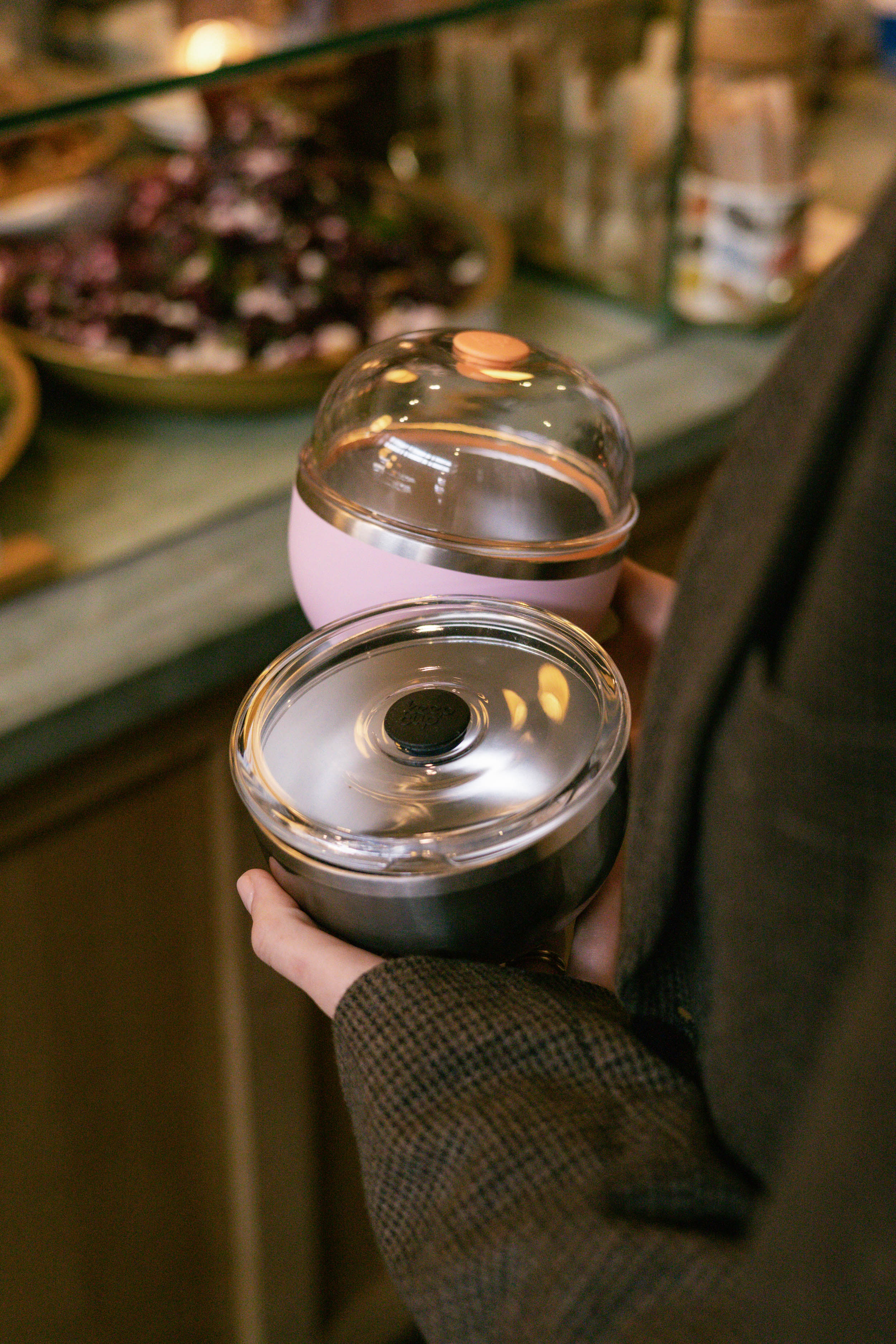 A hand holds two KeepCup Go Bowl products in a café setting - the Go Bowl Luxe in Pashmak Pink with a domed lid and peach button on top, alongside a Nitro bowl with a flat lid featuring a black center button.