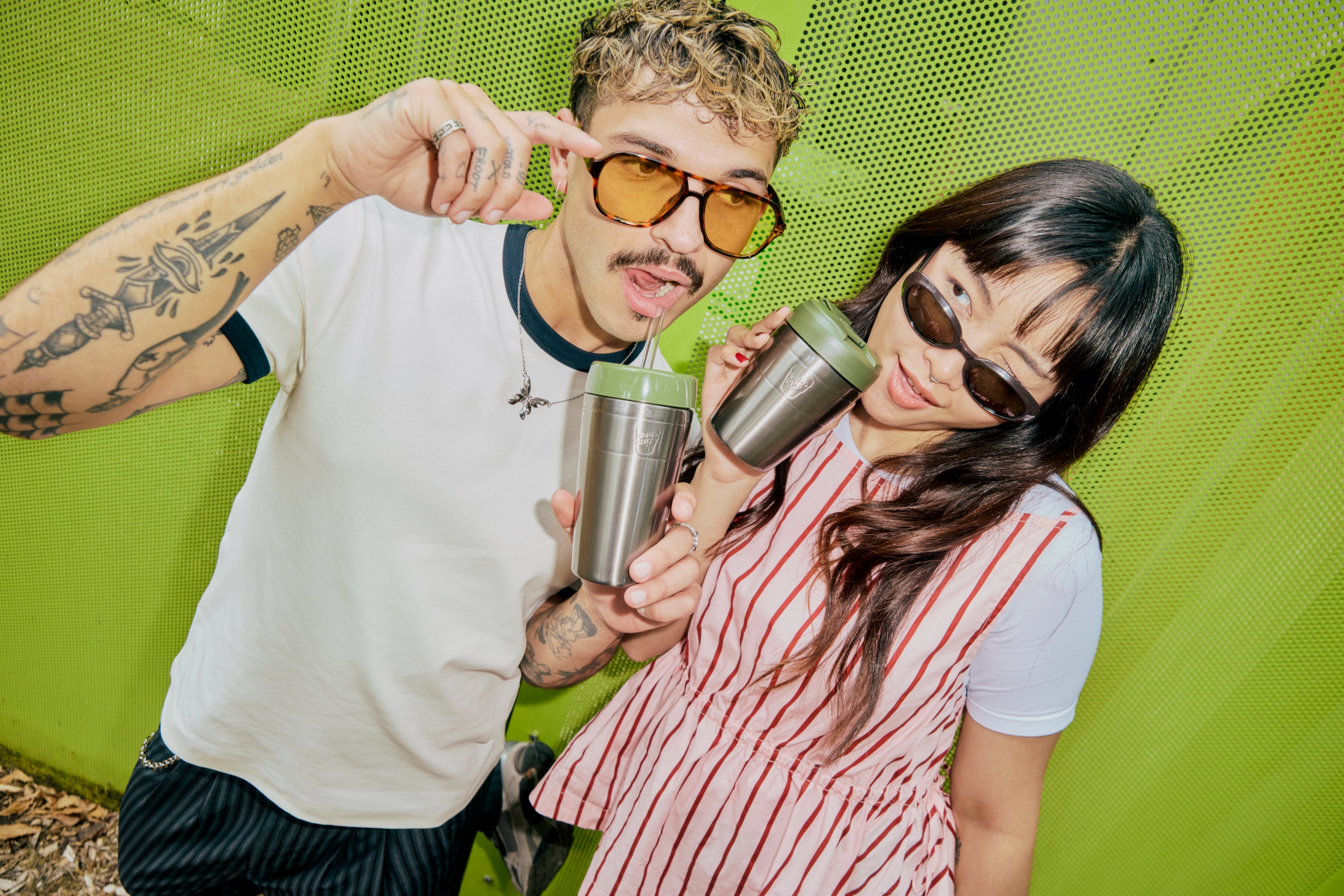 Two people with sunglasses drinking from stainless steel tumblers with green lids against a bright green dotted background