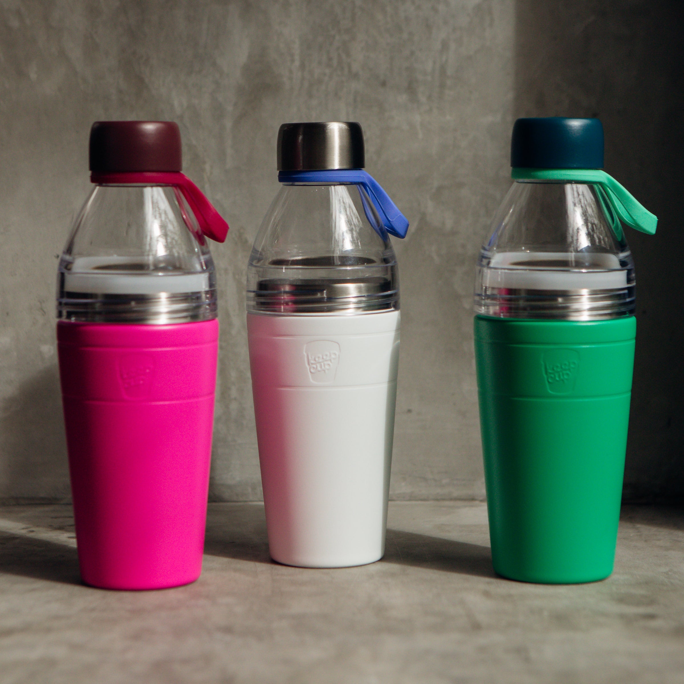 Three colorful KeepCup water bottles with clear top lids on a gray surface.