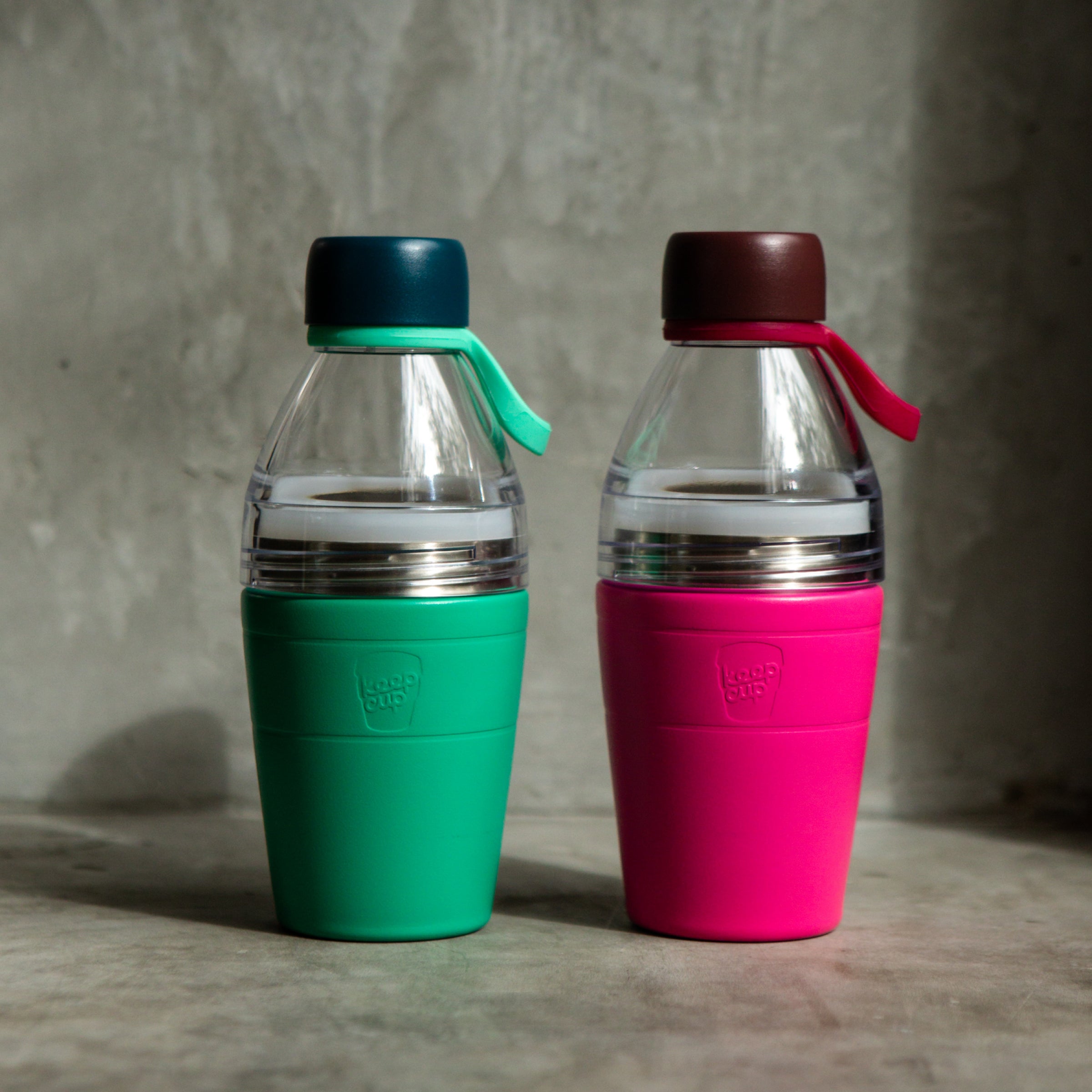 Two reusable KeepCup water bottles, one green and one pink, with clear top extensions on a textured surface.