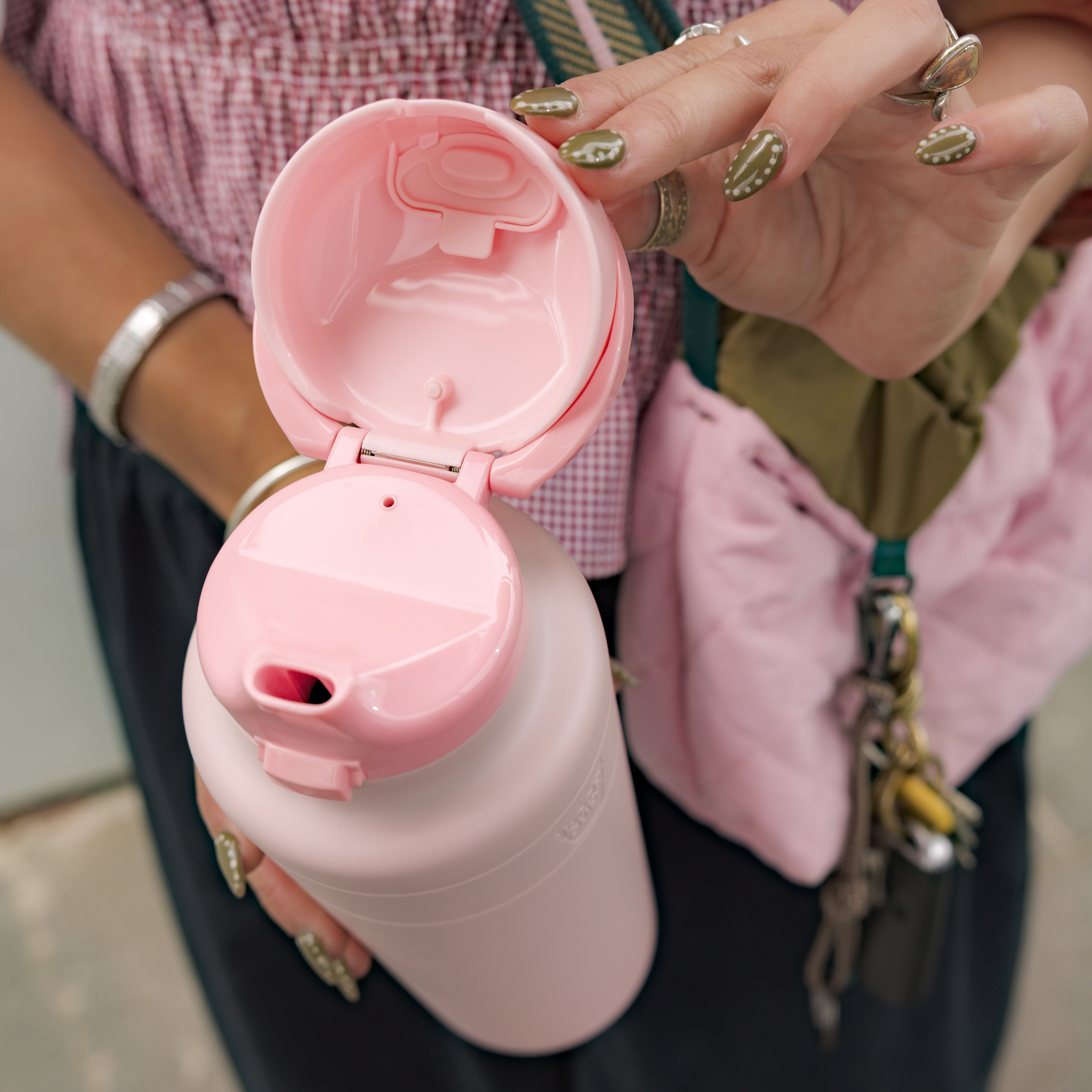 A woman's hand with olive green nail art and silver rings holds an open KeepCup Ora Bottle 24oz in Pashmak Pink, showing the flip-top lid and drinking spout, with a pink gingham blouse and olive accessories visible in the background.