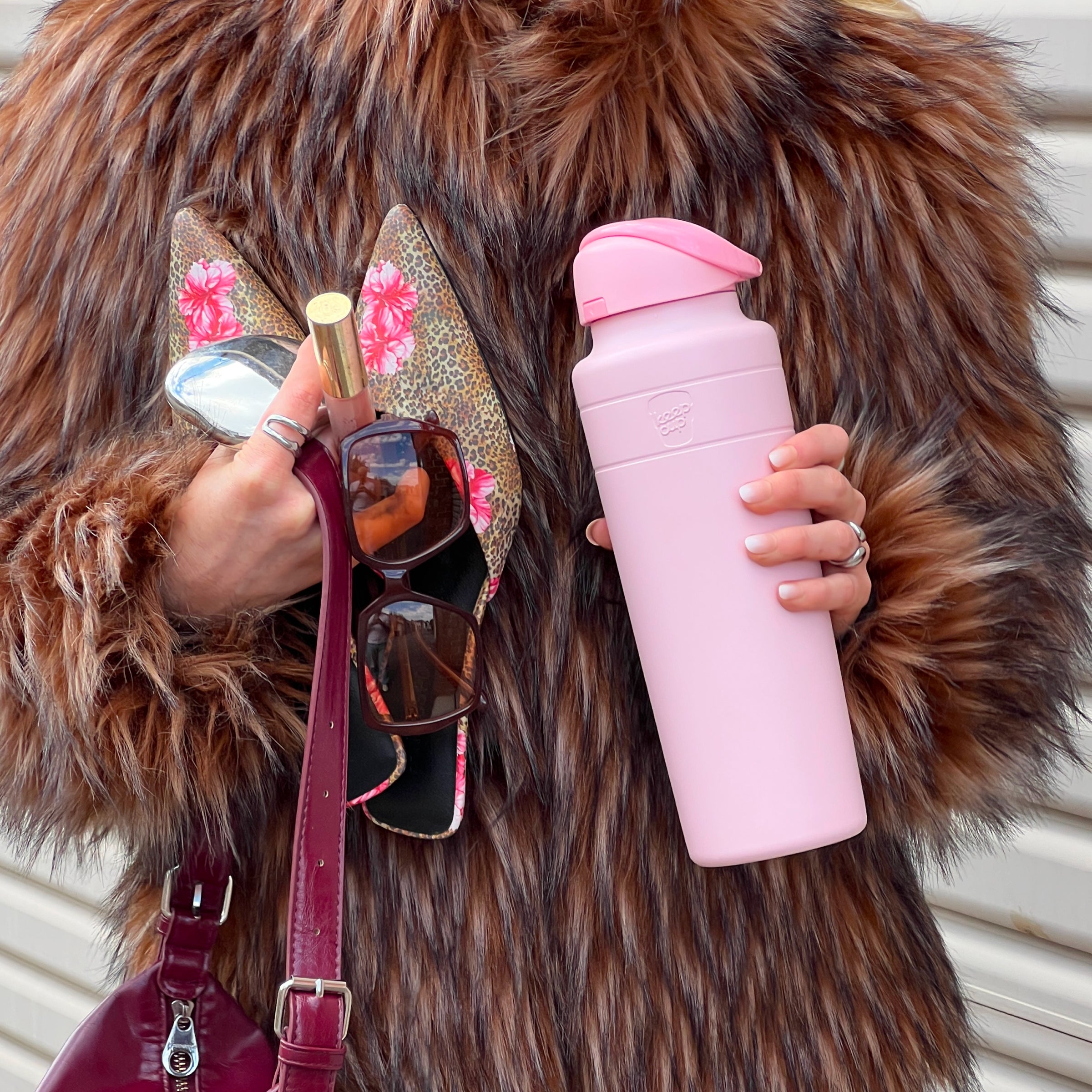 A person wearing a brown fur coat holds the KeepCup Ora Bottle 32oz in Pashmak Pink in one hand while the other hand holds sunglasses, leopard print heels with pink floral accents, and a gold lipstick tube, with a burgundy leather bag strap visible.