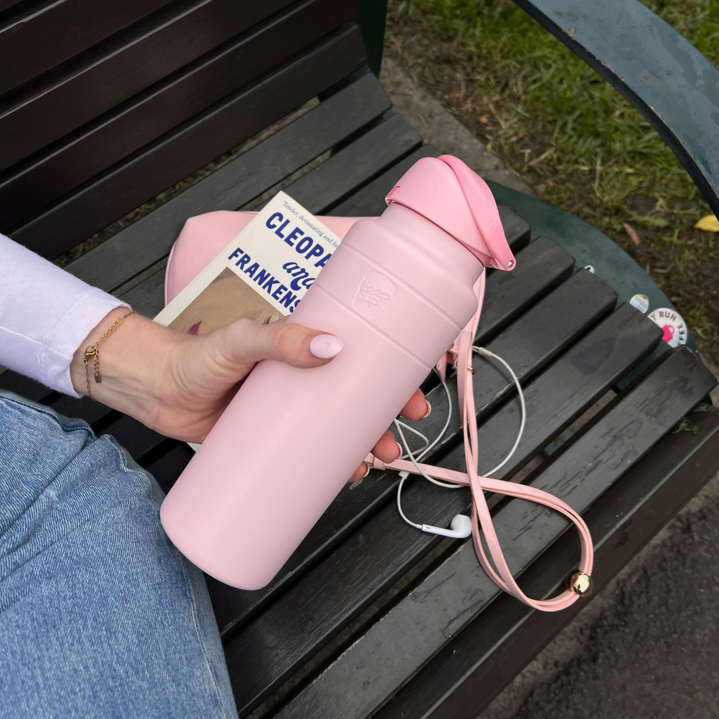 A hand with white nail polish holds the KeepCup Ora Bottle 32oz in Pashmak Pink while sitting on a dark wooden park bench, with a paperback book, white earbuds, and the bottle's pink carrying strap visible alongside.