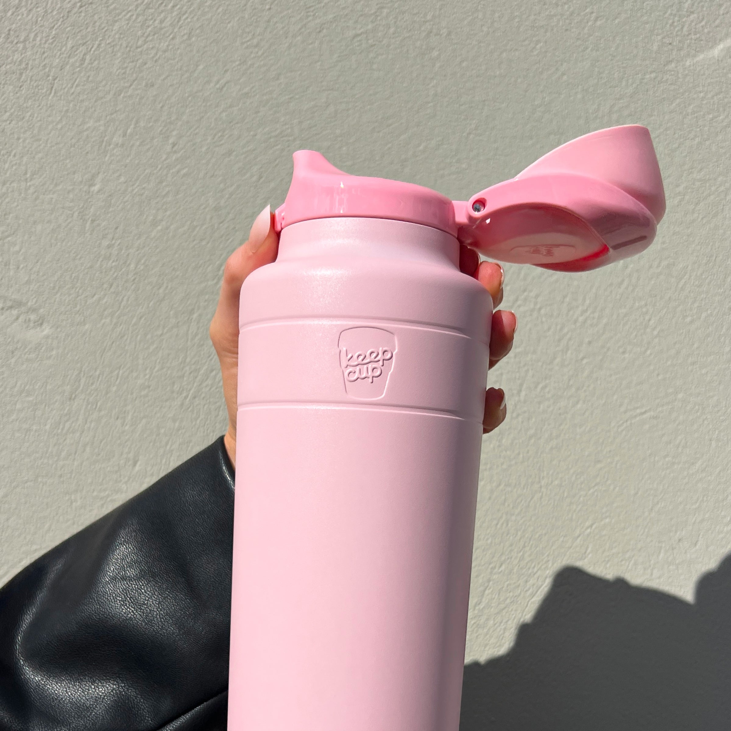 A hand with pink nail polish holds the Ora Bottle 32oz in Pashmak Pink against a light gray textured wall, with the flip-top lid open to reveal the drinking spout. The person wears a black leather jacket sleeve visible at the wrist.