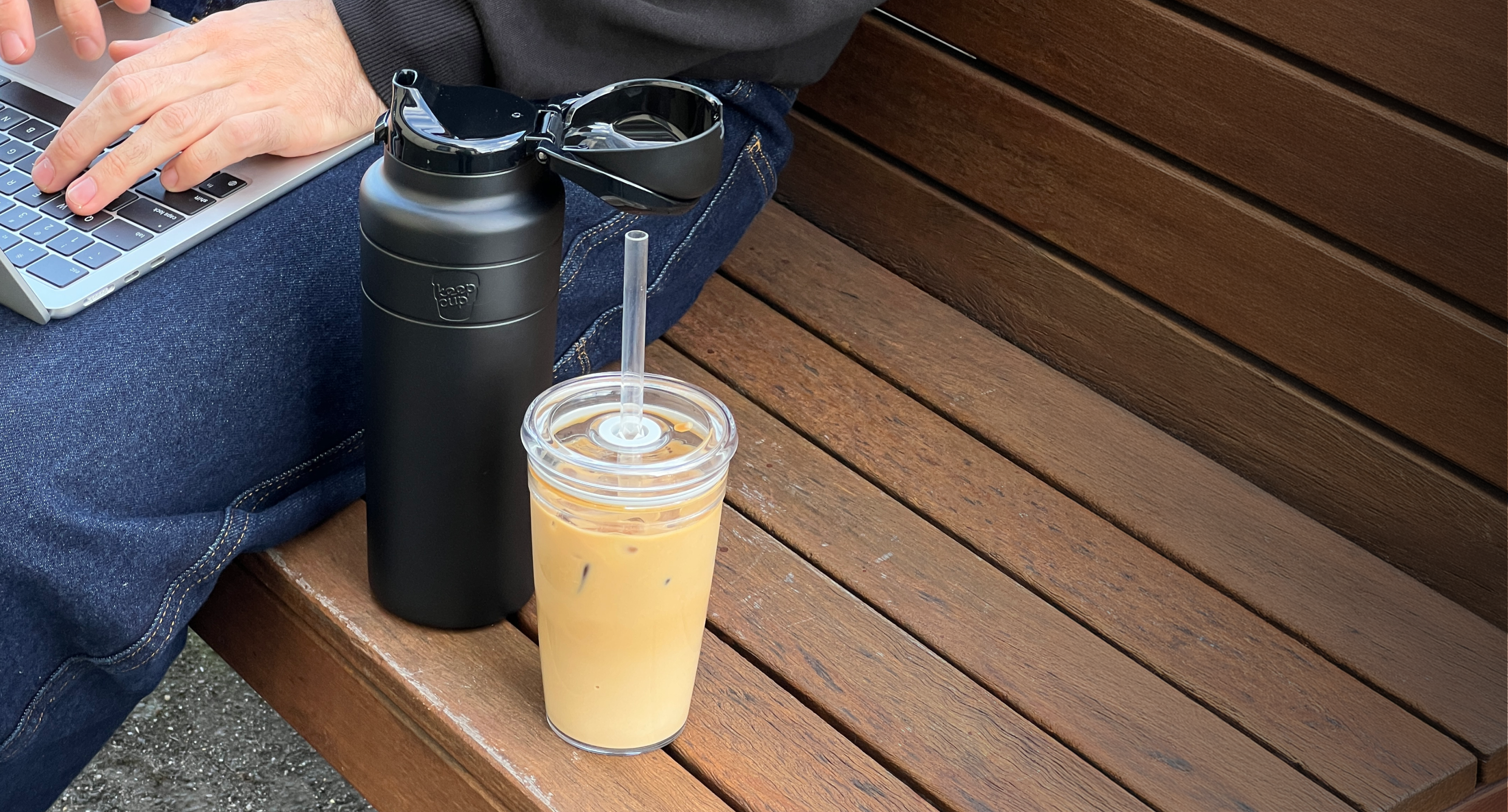 Person using laptop outdoors with black insulated water bottle and iced coffee on wooden bench, showing portable drinkware for active lifestyle