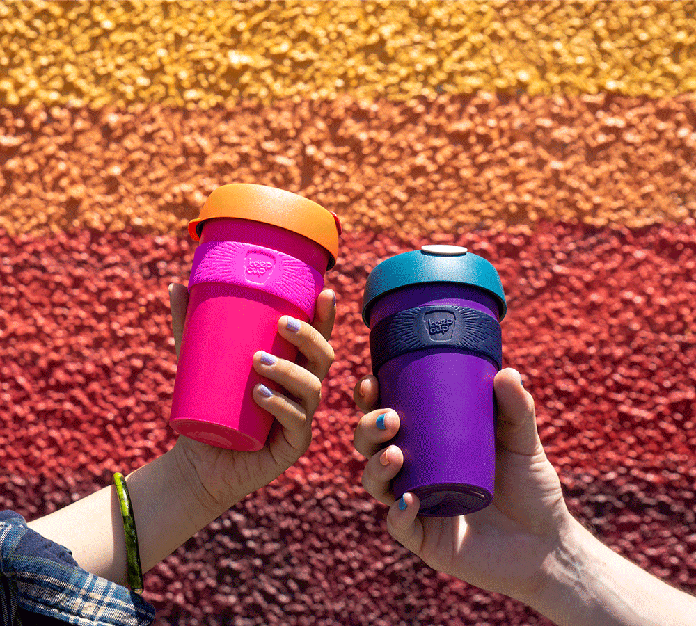 Two hands holding brightly coloured 12oz Original plastic reusable cups against a painted orange and yellow wall