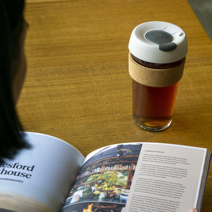 KeepCup Brew Cork Filter L 16oz filled with tea, featuring a latte lid with smoke plug and natural cork band, resting on a wooden table next to an open magazine about Daylesford Longhouse.