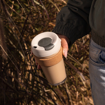 A hand holds the KeepCup Brew Cork L 16oz in Filter color at thigh level while walking through a natural outdoor setting with dried grass and twigs, showcasing the reusable cup's glass body, cork band sleeve, and latte lid with smoke plug