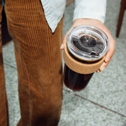 Hand holding a KeepCup Brew Cork Quicksip L 16oz filled with black coffee, showing the clear lid with sliding closure and natural cork band, against a terrazzo floor background.