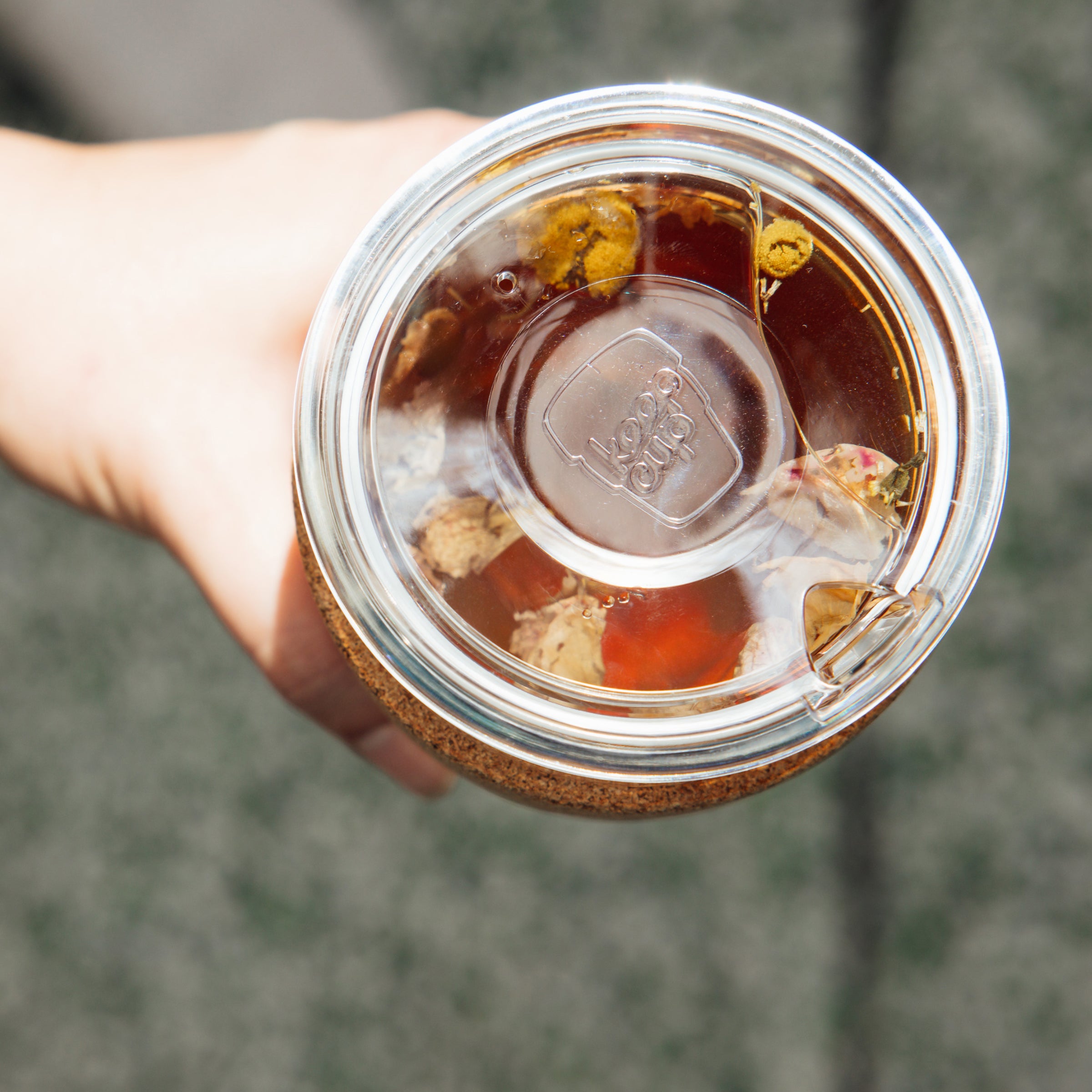 Hand holding a KeepCup Brew Cork Quicksip L 16oz viewed from above, showing the clear glass cup with cork band, transparent sipper lid, and loose leaf herbal tea with floating chamomile flowers steeping inside.