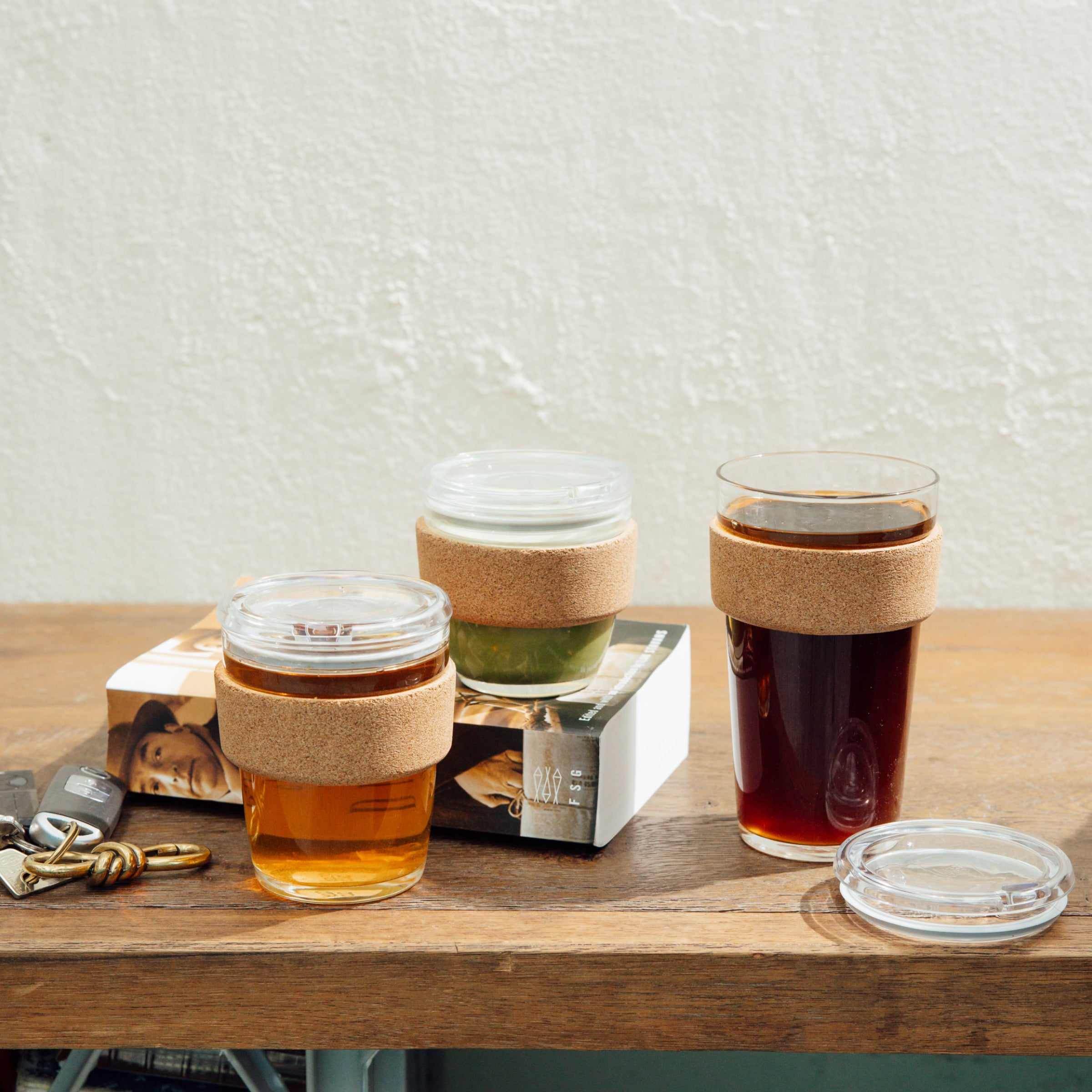 Three KeepCup Brew Cork cups in different sizes displayed on a wooden table, featuring the M 12oz Quicksip model with cork band and clear lid, shown alongside a book and keys in a casual lifestyle setting.