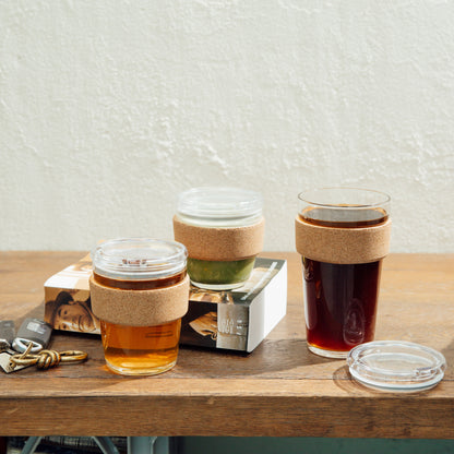 Three KeepCup Brew Cork cups in different sizes displayed on a wooden table, featuring the M 12oz Quicksip model with cork band and clear lid, shown alongside a book and keys in a casual lifestyle setting.