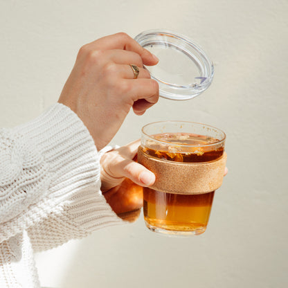 Hands holding a Brew Cork Quicksip M 12oz glass cup filled with golden tea, with one hand lifting the clear glass lid while the other grips the cork band sleeve, against a neutral background.
