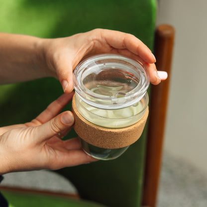 Hands holding a Brew Cork Quicksip S 8oz reusable glass coffee cup with cork band sleeve, filled with pale green tea, showing the clear lid from above while seated on a green velvet chair.