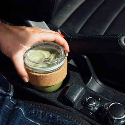 Hand reaching for a KeepCup Brew Cork Quicksip S 8oz in olive green, filled with a matcha latte, placed in a car cupholder next to the center console.