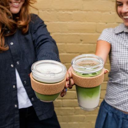 Two women smiling and holding up KeepCup Brew Cork Quicksip cups filled with layered matcha lattes, featuring glass construction with cork band sleeves and clear Quicksip lids, against a yellow brick wall background.