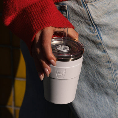 White KeepCup reusable coffee cup with clear lid and metal straw held by person in red sweater and jeans