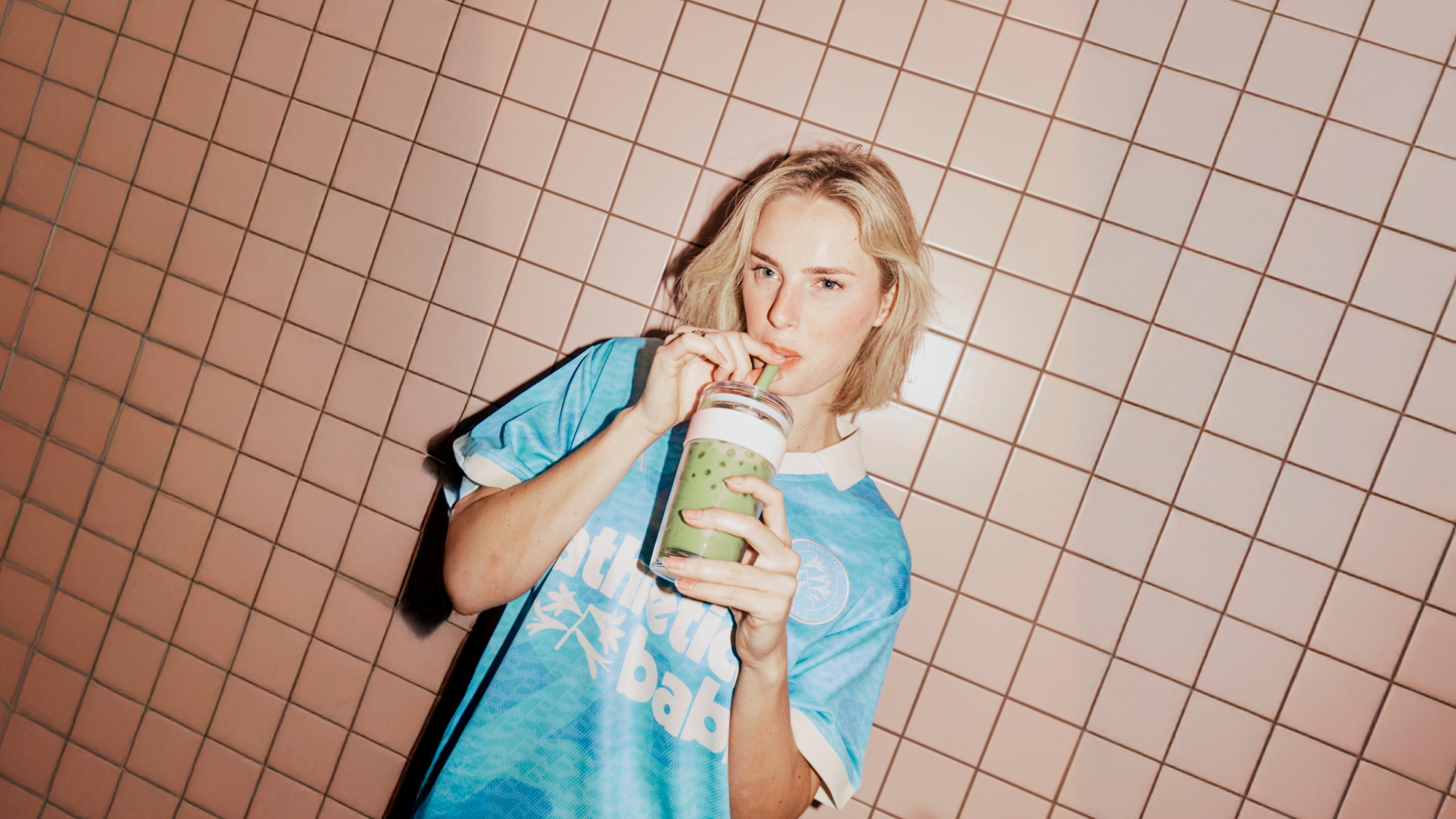 Person in a blue shirt holding a green smoothie in a tiled room