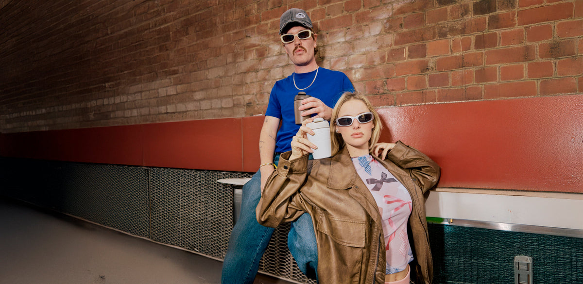 Two people wearing trendy sunglasses in casual setting - man in blue shirt with gray cap, woman in brown leather jacket holding white cup
