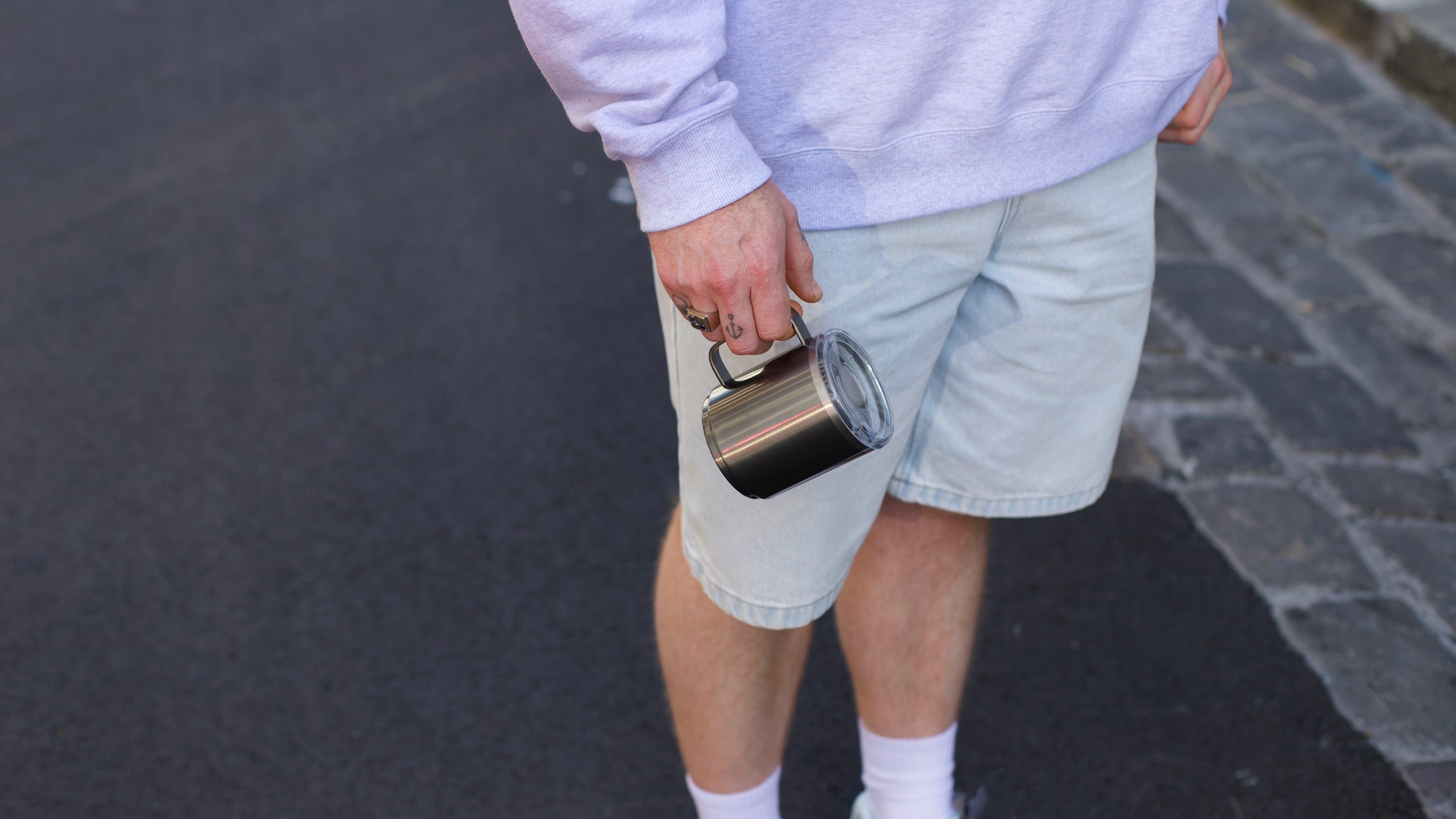 Person holding stainless steel insulated tumbler with clear lid, wearing white sweatshirt and gray shorts outdoors