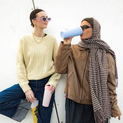 Two stylish women leaning against a white wall, one wearing a cream cable-knit sweater holding a KeepCup Commuter XL 20oz in Pashmak Pink, while her companion in a brown leather jacket and houndstooth headscarf drinks from a Orchid KeepCup.