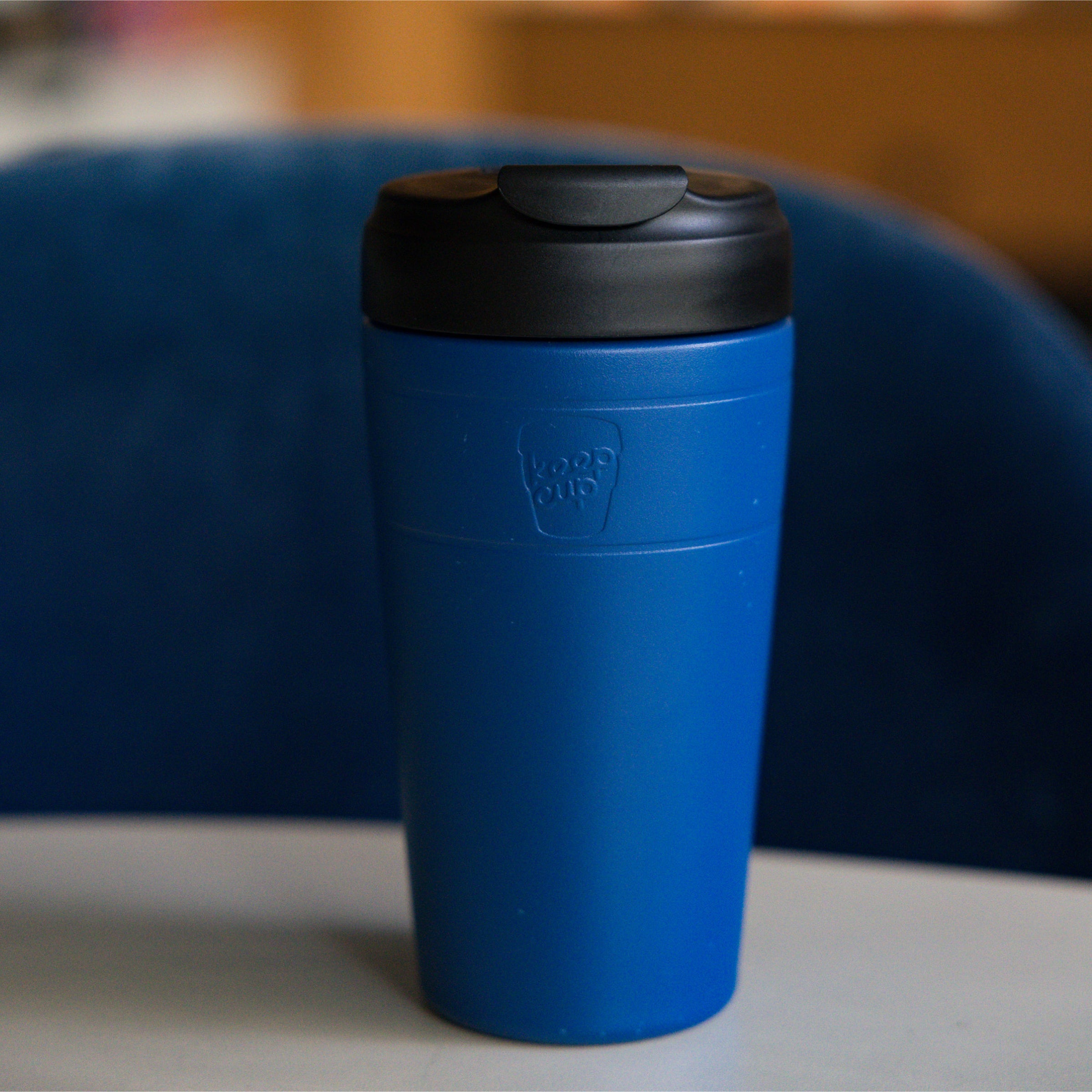KeepCup Commuter L 16oz in Cobalt Black, featuring a cobalt cup with black lid, displayed on a gray surface against a blue upholstered chair in a lifestyle setting.