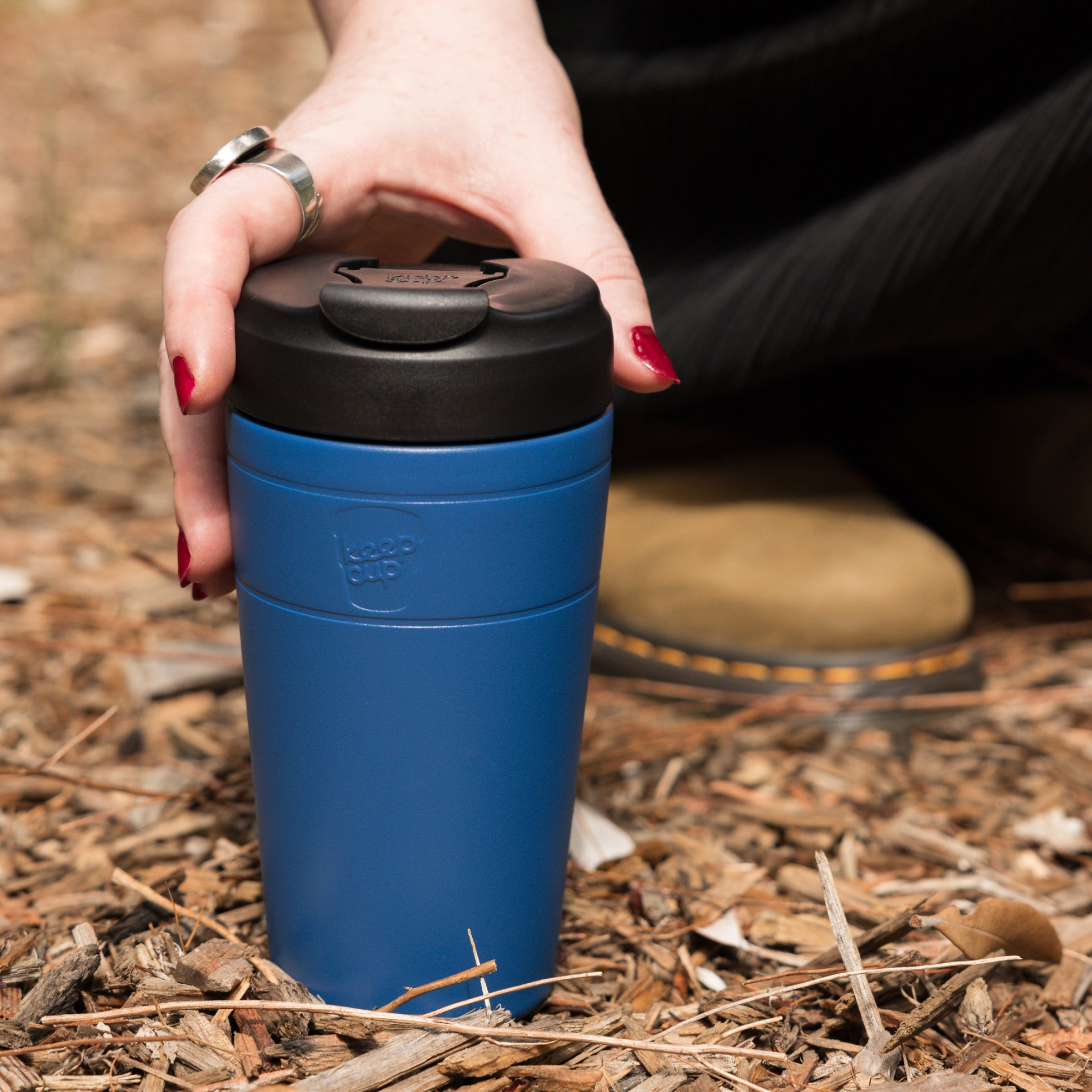 A hand with red nail polish reaches down to grasp a KeepCup Commuter L 16oz in Cobalt Black, resting on wood chip mulch outdoors, with the person's boots visible in the background.