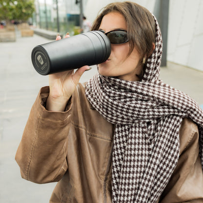 Woman wearing sunglasses, a brown leather jacket, and a houndstooth scarf drinks from a KeepCup Commuter XL 20oz in Black while walking outdoors in an urban setting.