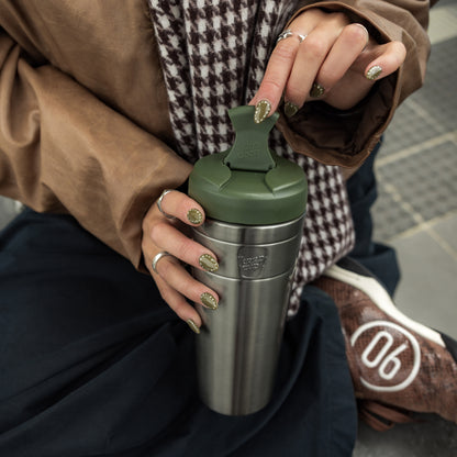 Woman holding stainless steel water bottle with olive green lid, wearing matching green nail polish and brown coat