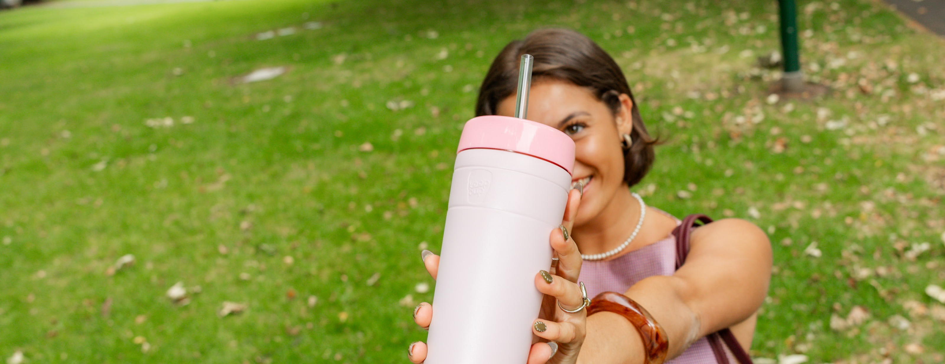 Woman holding pink insulated tumbler with straw outdoors, wearing pearl necklace and purple top in park setting