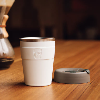 White KeepCup reusable coffee cup with gray lid sitting on wooden table in cozy cafe setting
