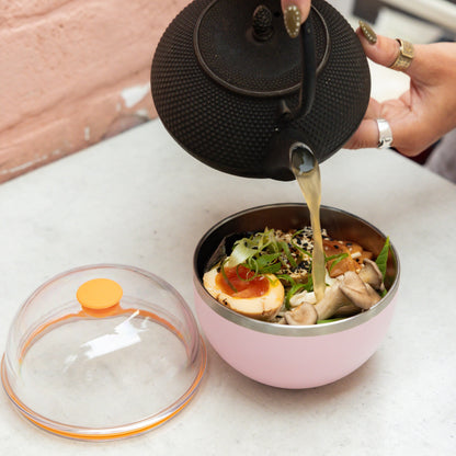 Person pouring tea from a black teapot into a pink bowl with food on a white surface.