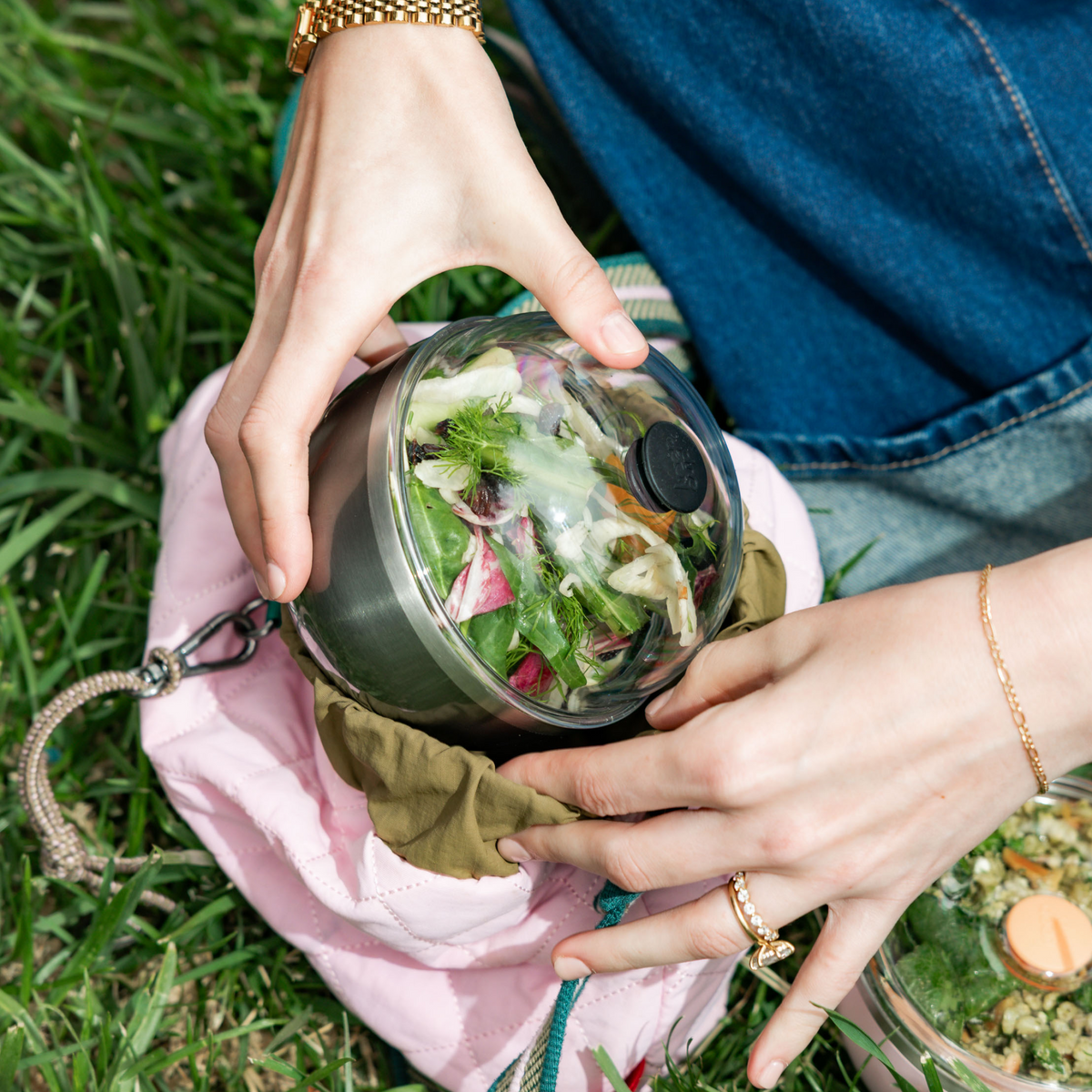 Hands holding a KeepCup Go High Go Low in Nitro filled with a fresh salad, being placed into a pink quilted bag on grass during an outdoor picnic setting.