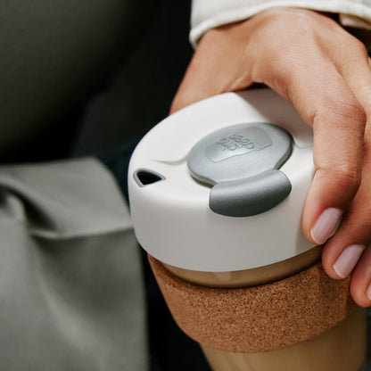 A hand holding a KeepCup Brew Cork L 16oz in Filter colourway, viewed from above showing the white lid with grey plug and sip hole, with the natural cork band visible around the glass cup.