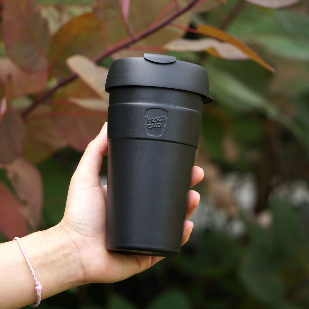 Black reusable coffee cup with lid held in hand against blurred green foliage background